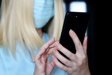 Blonde woman in face mask using smartphone, mobile phone in female hands closeup. Concept of safety work in office during coronavirus pandemic