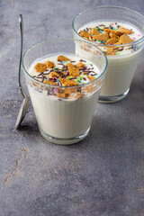 Top view of two glasses with natural yogurt, cookie pieces and colors with spoon, on gray background, in vertical