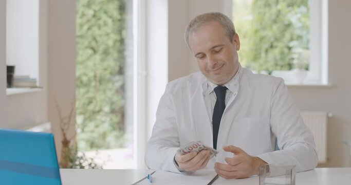 Portrait of satisfied doctor counting and smelling money indoors. Smiling Caucasian man sitting in hospital office with dollars. Rich physician enjoying his job. Bribery concept. Cinema 4k ProRes HQ.