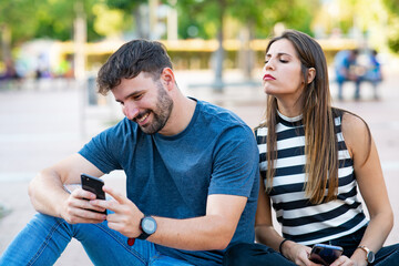Cheating man chatting with women on his smartphone while his girlfriend spies on him sitting on a...