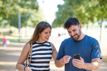Man shows happy girl his mobile phone pointing at screen with finger in park