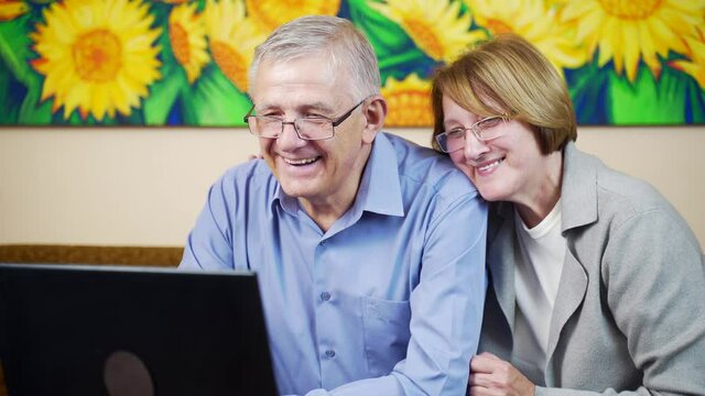 Close up portrait of cute seniors old couple sitting in eyeglasess using computer then looking at each other in living room, painting background. view video photo memories. Happy smiling and hugging