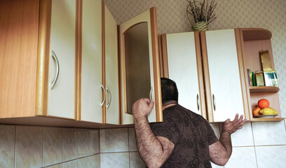 Young man in shirt peeking  into kitchen cupboard at home, viewed from the back side. alone hungry...
