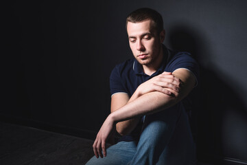 Young attractive man in blue jeans and a polo shirt sits on the floor near the wall in the studio. Youth and attractiveness concept
