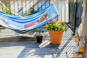 woman in stripe top sunbathing peacefully on terrace or balcony hammock. Attractive lady relax mode on summer holidays. Girl sunbathing under bright scorching sun on hammock, flower pot near.
