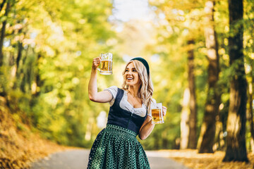 Pretty happy blonde in dirndl, traditional festival dress, holding two mugs of beer outdoors in the forest