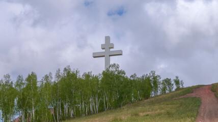 Large white cross on the hill. Dirt road up. Green trees, sky with clouds. Calvary concept, path to the crucifixion.