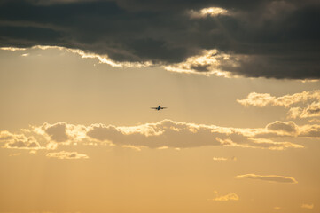 Beautiful dramatic gray cloud on orange sunset sky. Small silhouette of an airplane in the distance rising upward the clouds after takeoff. Air transport, natural colorful pattern sky background.