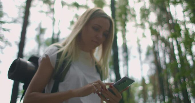 A young woman with a mobile phone walks through the forest traveling with a backpack in slow motion. Navigate through the forest using the Navigator in your mobile phone