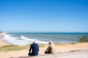 Fototapeta premium Back view of old men sitting on the bench near ocean, kiteboarding or kitesurfing on sunny outdoors background.