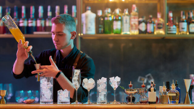 Young male bartender pouring, mixing ingredients while making classic cocktail alcoholic drink at the bar counter in the night club
