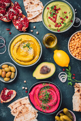 Ingredients for yellow turmeric hummus, green parsley hummus and red beetroot hummus on dark background, lemon, sesame, olive oil, pita, avocado, pomegranate. Middle eastern, jewish, arabic cuisine.