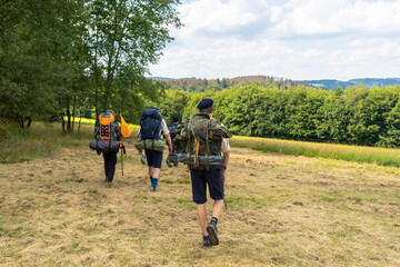 Scouts or tourists go with big backpacks on the green forest trail on sunny summer day. Active healthy lifestyle people with rucksacks. Hiking Trekking Scouting. Green forest background. - obrazy, fototapety, plakaty