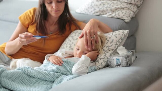 Sick child, toddler boy lying on the couch with his white puppy in living room with fever, resting at home