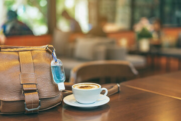 Mini alcohol gel bottle to kill Corona Virus(Covid-19) hang on a brown leather shoulder bag with hot coffee on table in cafe.New normal lifestyle.Health care concept.Selective focus on alcohol gel
