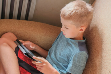 Blonde caucasian boy playing video games/watching videos on tablet close-up. Gadget concept. Kids and technologies