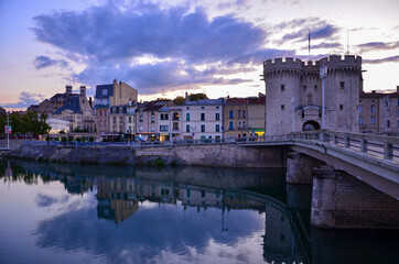 Obraz premium Cityscape of the historic town of Verdun in the Northeast of France before sunset, river Meuse (Maas) in front, medieval toll gate on the right, cloudy evening sky in summer, panoramic view