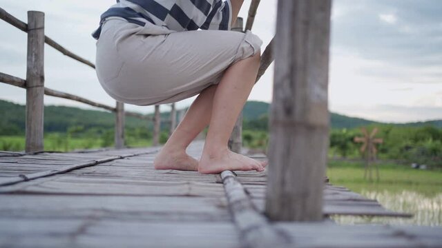 Slow motion Young female sitting down legs hanging on the edge of wood bridge, relaxation swing legs, Asian country side culture traditional rice growing field, calm relaxation, lower body section