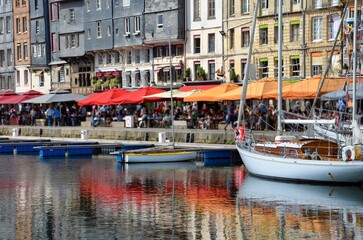 Fototapeta na wymiar The old port(Vieux Bassin)of the town of Honfleur in the county of Calvados in Normandy,France,traditional historic houses in the background, people under parasols at the waterfront, boats on the quay