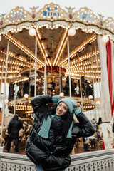 Fototapeta na wymiar Beautiful smiling woman posing near carousel in Christmas market. Outdoor photo of happy dark-haired girl relaxing in amusement park in winter. Street style.