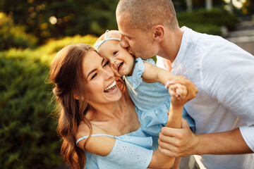 Overjoyed family walking at the park, loving mom hold smiling toddler in arms, laughing, caring dad kissing little daughter at the cheek, happy family moments, parenthood concept