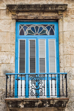 Close-up of an old blue wooden window with white shutters and a small balcony with a decorative black metal grid, in Old Havana.