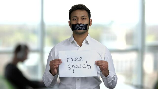 Dark-skinned man with black tape over mouth. Indian man holding paper card with inscription Free speech. Abstract blurred background.