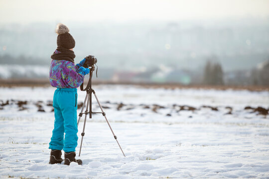 Child boy taking pictures outside in winter with photo camera on a tripod on snow covered field.