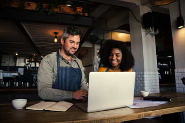 Male and mixed race female coffee shop owners smiling at laptop in coffee shop.
