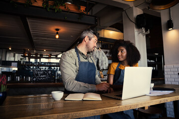 Male manager advising waitress in apron behind laptop and note book while standing in trendy coffee shop.