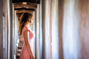 Young woman in pink dress side profile standing looking through window of wall of fortress tunnel passage in Castiglione del Lago in Italy