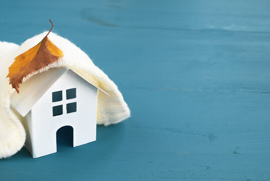 White model house with warm clothes and autumn leaf on wooden blue table. Heating system, cold season concept. Copy space, close up