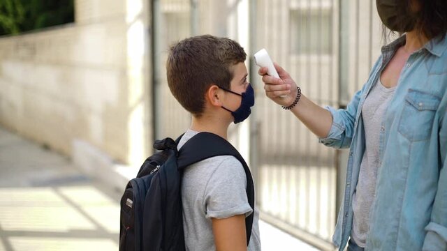 A Queue Students Checking Body Temperature By Infrared Thermometer In Front Of School On Daytime. Elementary Pupils Wearing Face Masks. Concept Back To School, Coronavirus And Social Distance.