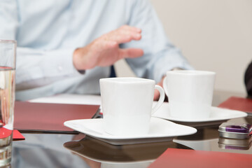 glass table with white coffee cups and plates, next to documents, in the background a person in a shirt