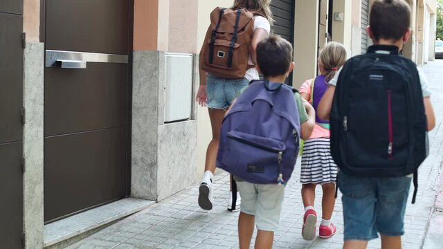 The backs of four children walking down the street, going to school. It's the first day of school during coronavirus pandemic. They wear protective face masks and backpacks. Rear view.