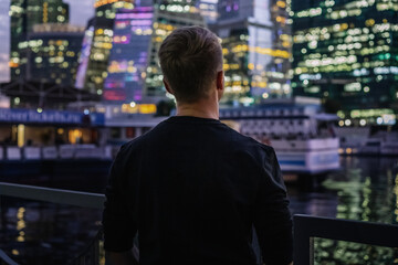 A business man in a black sweater stands with his back to the camera with a view of the night illumination business skyscrapers in Moscow