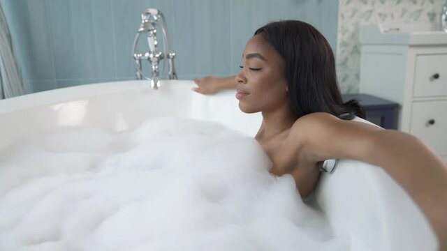 African American woman lying in bubble bath and looking at window. Wellness and relaxation concept. Panning camera