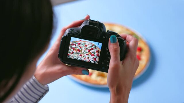  Food blogger of blogging concept. Top view - Female hands use dslr camera photographing appetizing fresh cooked italian pizza with tomatoes, cheese mozzarella, and sauce pesto on blue background. 