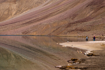 Two tourists - a photographer and his model walk on the bank of chandratal - a high altitude lake with clear water and textured colorful hill slope in background and its reflection on a clear day