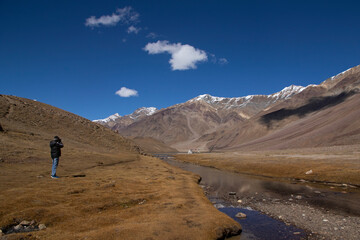 A tourist photographer near Chandratal uses his camera to click photos of scenery - the blue sky with white clouds, shadow of clouds, reflection on water, snowy peaks and yellow grass.