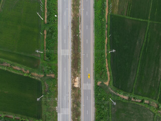 Aerial photograph of a yellow car running on a paved road that cuts through the green fields.