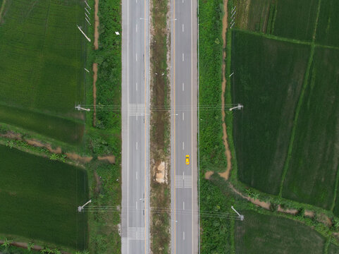 Aerial Photograph Of A Yellow Car Running On A Paved Road That Cuts Through The Green Fields.