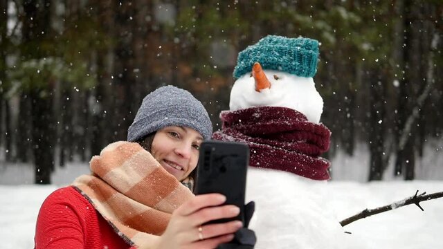 One happy winter day, a young girl is photographed near a snowman in a green knitted hat and scarf. Winter, new year, falling snow, christmas.