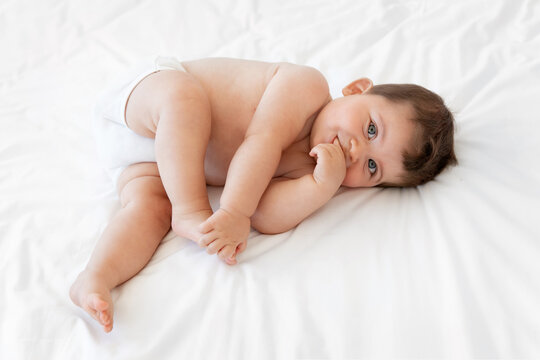 Chubby baby lying on white bed sucking finger and holding foot