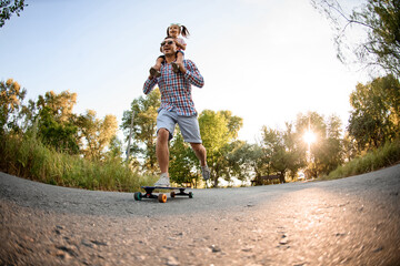 cheerful young man holds little girl on his shoulders and ride skateboard in the park