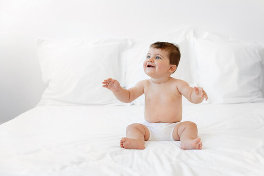 Happy chubby baby sitting on white bed raising arms