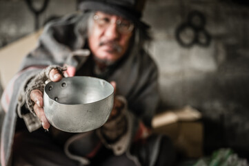 Homeless old man with gray beard and hair sitting holding metal bowl with food beggar because...