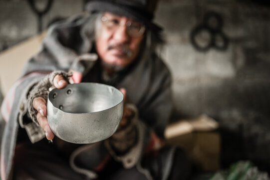 Homeless old man with gray beard and hair sitting holding metal bowl with food beggar because hungry and cold on walkway street. Poor senior man homeless and depression concept.