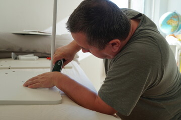 Man assembling furniture with power cordless screwdriver. He is redecorating his home with new computer desk. The DIY set was ordered on an online store and delivered by mail.