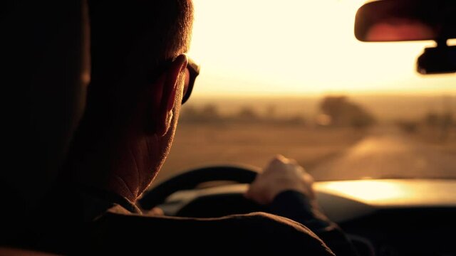 Male businessman drives a car while traveling. Man's hands lie on the steering wheel in the rays of the sunset. Silhouette of a man driving a car. Business, travel concept.